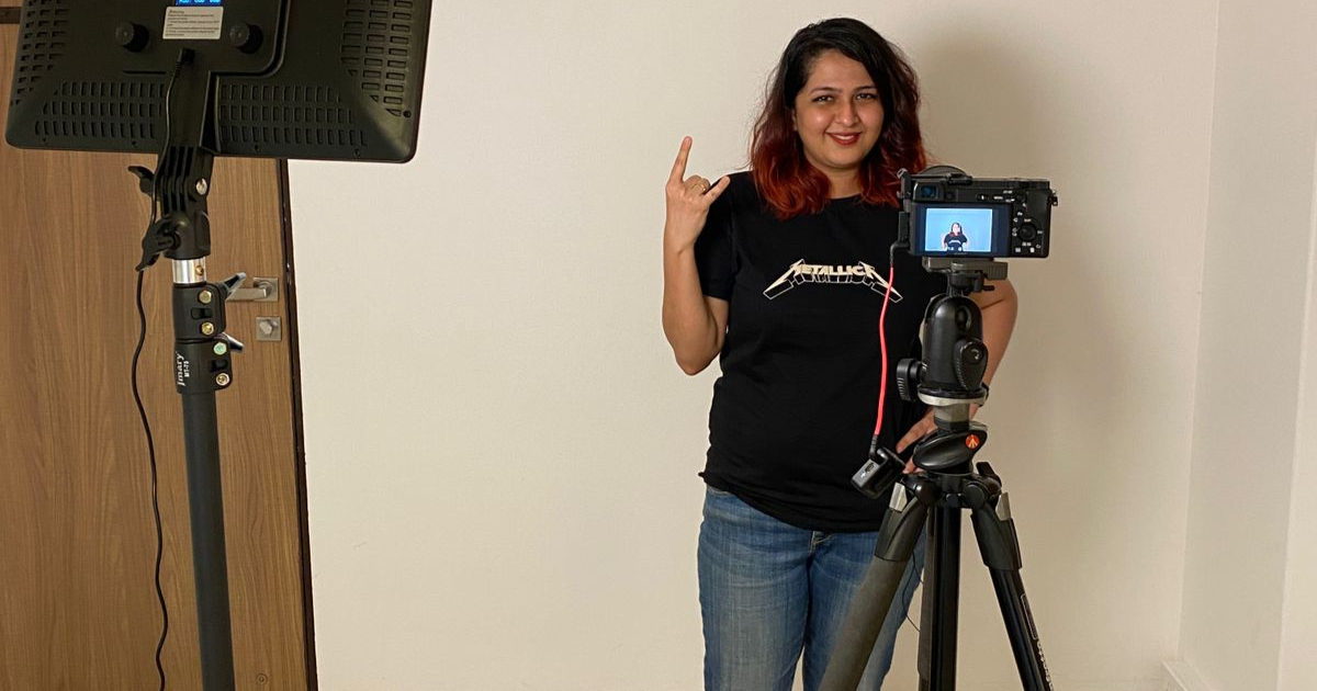 A woman stands in front of a camera on a tripod. She's holding up the metal sign. Her hair is slightly red in color and she's wearing a Metallica tshirt. The preview pane of the camera is on and you can see her in it too