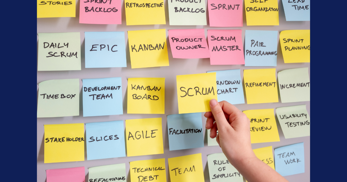 A physical Scrum board with multiple sticky notes of various colors. They have words like Agile, slices, stakeholders, team, Kanban, Epic, Timebox, etc. written on them. A hand picks out a yellow sticky note that has Scrum written on it