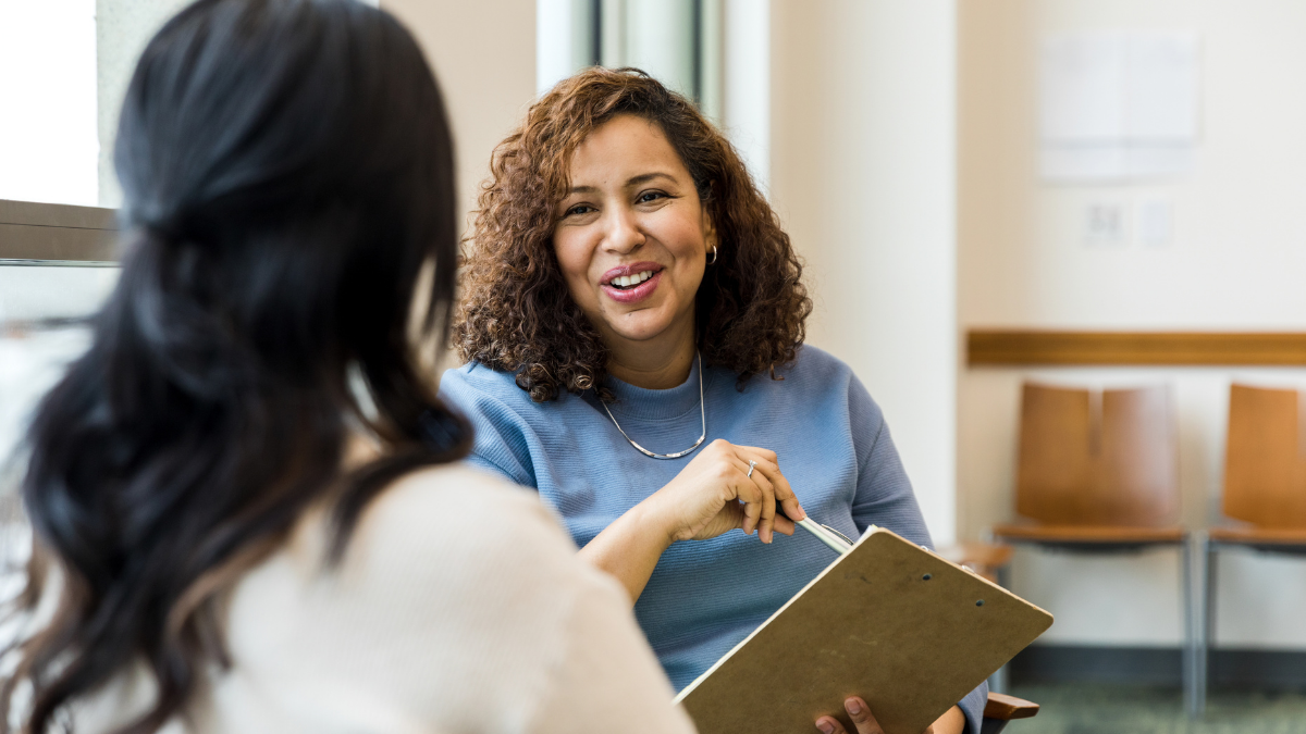 A woman with brown, curly hair speaking to a woman whose back is against the camera. The brown haired lady has a notepad and pen in her hand and she's smiling